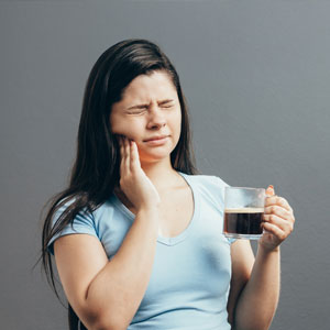 Young woman with sensitive teeth and cup of hot coffee on gray background